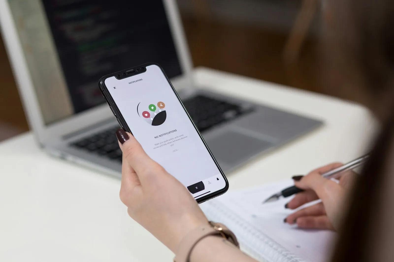 A person checking an app on their phone while taking notes, with code open on a laptop behind them